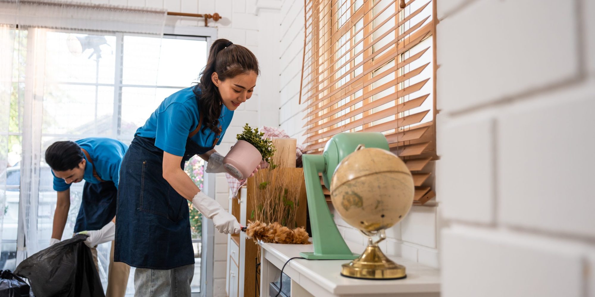 Asian young man and woman cleaning service worker work in living room. Attractive housekeeper cleaner team wear apron and cleaning messy dirty floor for housekeeping housework and chores in house.